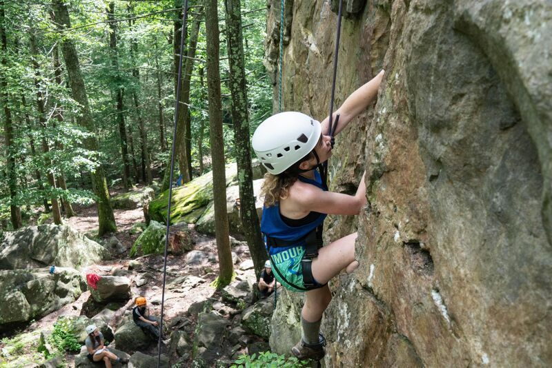 A woman wearing a white helmet is rock climbing on a steep rock face. She is secured with a rope. The rock face is textured and rugged. In the background, there is a forest with tall trees and a path. Other people are watching the climber. The scene is outdoors, likely on a sunny day.
